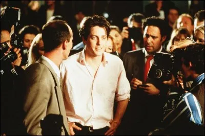 En chemise rose dans la foule, Hugh Grant regarde droit devant lui en souriant. Que ou qui regarde-t-il ?